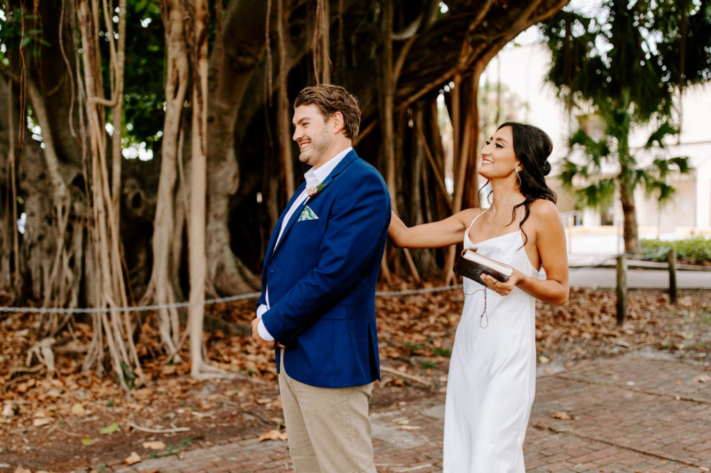 Tampa Bay Bride and Groom First Look under St. Petersburg Banyan Trees, Bride Wearing White Show Me Your MuMu Wedding Dress with Boho Inspired Hair and Makeup, Groom wearing Navy Suit Jacket with Casual Beige Bottoms and Tropical Handkerchief | Florida Wedding Planner Parties A'La Carte