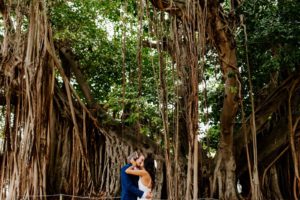 Tampa Bay Bride and Groom Intimate Embrace Kiss under St. Petersburg Banyan Trees, Bride Wearing White Show Me Your MuMu Wedding Dress with Boho Inspired Hair and Makeup, Groom wearing Navy Suit Jacket | Florida Wedding Planner Parties A'La Carte