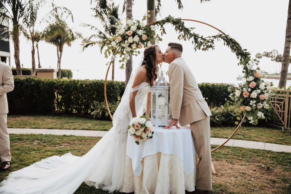 Earthy and Elegant Wedding Ceremony Decor, Bride and Groom Exchanging Kiss During Ceremony, Metal Circular Arch with Lush Greenery, Blush Pink and White Flowers | Tampa Bay Wedding Dress Shop Truly Forever Bridal