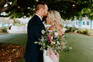 Tampa Bay Bride Holding Wild Floral Bouquet with Greenery Eucalyptus, Pink and Purple Flowers, Groom Kissing Bride's Forehead