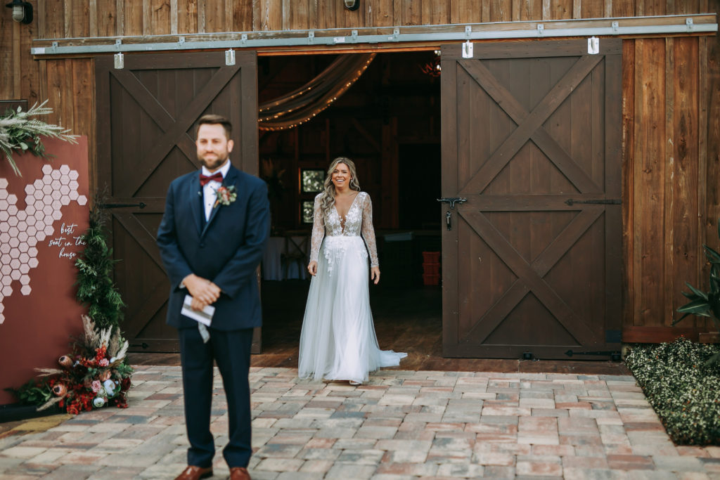 Rustic Bride In Romantic Long Sleeve Lace and Illusion Plunging V Neckline Flowy Skirt Wedding Dress Walking To Husband for First Look, Husband Turned with Back to bride in Navy Blue Tuxedo and Burgundy Bow Tie | Tampa Bay Wedding Venue Mision Lago Estate