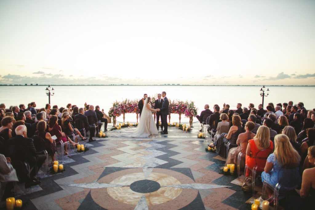 Bride and Groom Exchanging Vows during Outdoor Waterfront Tampa Bay Sarasota Wedding Ceremony with Ground Arch Floral Arrangements featuring Colorful Pink Purple and Orange Roses with Candles