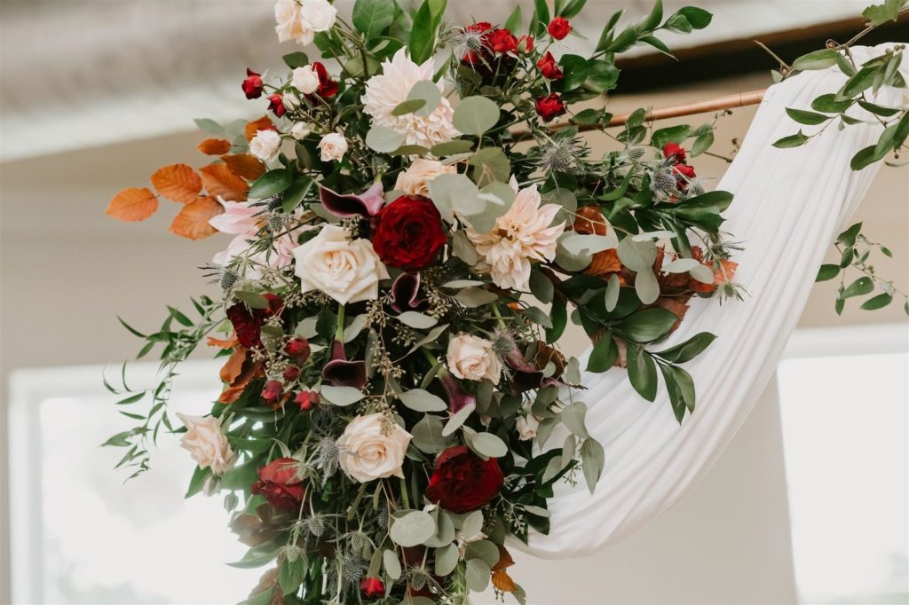 Draped Wedding Ceremony Arch with Fall Greenery Floral Arrangement Spray of Copper Leaves, White and Red Roses, Zinnia, and Eucalyptus | Botanica International Design