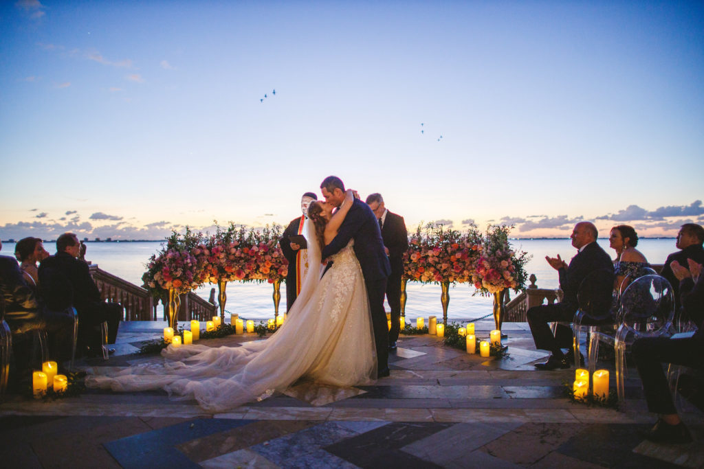 Bride and Groom First Kiss during Outdoor Waterfront Sunset Tampa Bay Sarasota Wedding Ceremony with Ground Arch Floral Arrangements featuring Colorful Pink Purple and Orange Roses with Candles