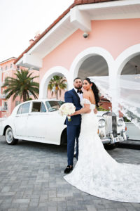 Florida Bride and Groom with Veil Flowing In Wind In Front of White Vintage Rolls Royce, Bride Wearing Rivini Fit and Flare Lace Wedding Dress, Holding Classic Round Bouquet with Ivory and Blush Pink Roses | Historic Tampa Bay Wedding Venue The Don CeSar | Florida Wedding Photographer Limelight Photography | St. Pete Beach Wedding Florist Iza's Flowers | South Tampa Bridal Boutique Isabel O'Neil Bridal Collection