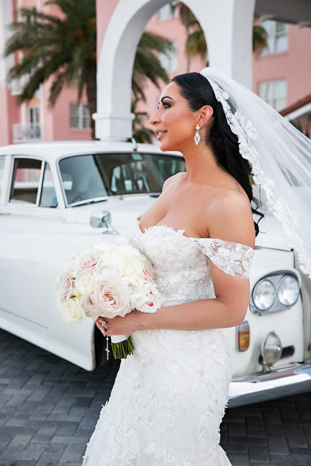 Florida Bride with Veil Flowing In Wind In Front of White Vintage Rolls Royce, Bride Wearing Rivini Off The Shoulder Fit and Flare Lace Wedding Dress, Holding Classic Round Bouquet with Ivory and Blush Pink Roses | Historic Tampa Bay Wedding Venue The Don CeSar | Florida Wedding Photographer Limelight Photography | St. Pete Beach Wedding Florist Iza's Flowers | South Tampa Bridal Boutique Isabel O'Neil Bridal Collection
