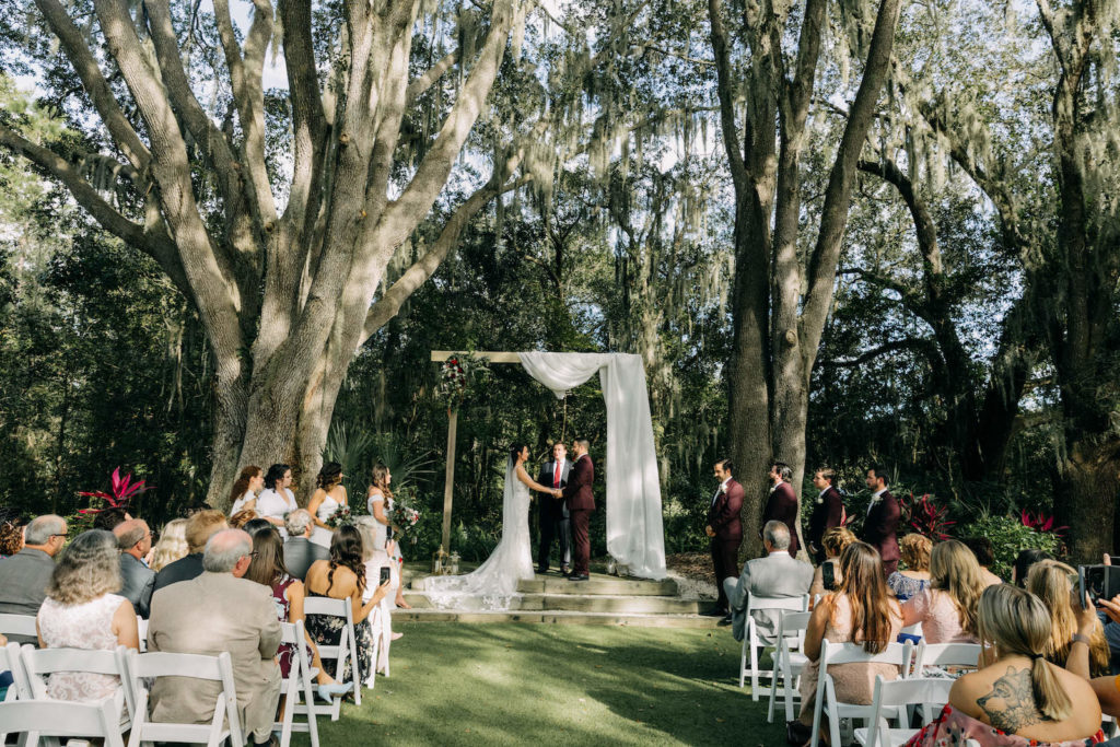 Bride and Groom Outdoor Garden Wedding Ceremony Exchanging Wedding Vows Under Rectangular Wooden Arch with White Linen Drapery Under Trees | Tampa Bay Wedding Photographer Amber McWhorter Photography | Wedding Venue The Secret Garden at Paradise Spring
