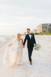 Romantic Florida Bride in Lace with Nude Lining Wedding Dress Holding Hands with Groom with Navy Blue Suit Holding Eucalyptus Greenery and White Floral Bouquet Walking Holding Hands on Beach | Sarasota Wedding Venue The Resort at Longboat Key Club