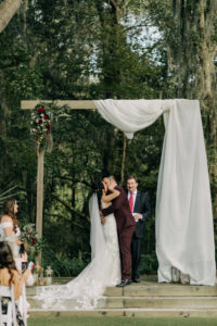 Bride and Groom Outdoor Garden Wedding Ceremony Exchanging Wedding Vows Under Rectangular Wooden Arch with White Linen Drapery Under Trees | Tampa Bay Wedding Photographer Amber McWhorter Photography | Wedding Venue Paradise Spring
