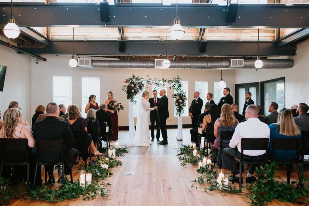 Bride and Groom Exchanging Vows During Intimate Indoor Historic Industrial Loft Tampa Wedding Ceremony with Black Chairs | Draped Ceremony Arch with Fall Greenery Floral Arrangement Spray and Greenery Vine Aisle with Pillar Candles