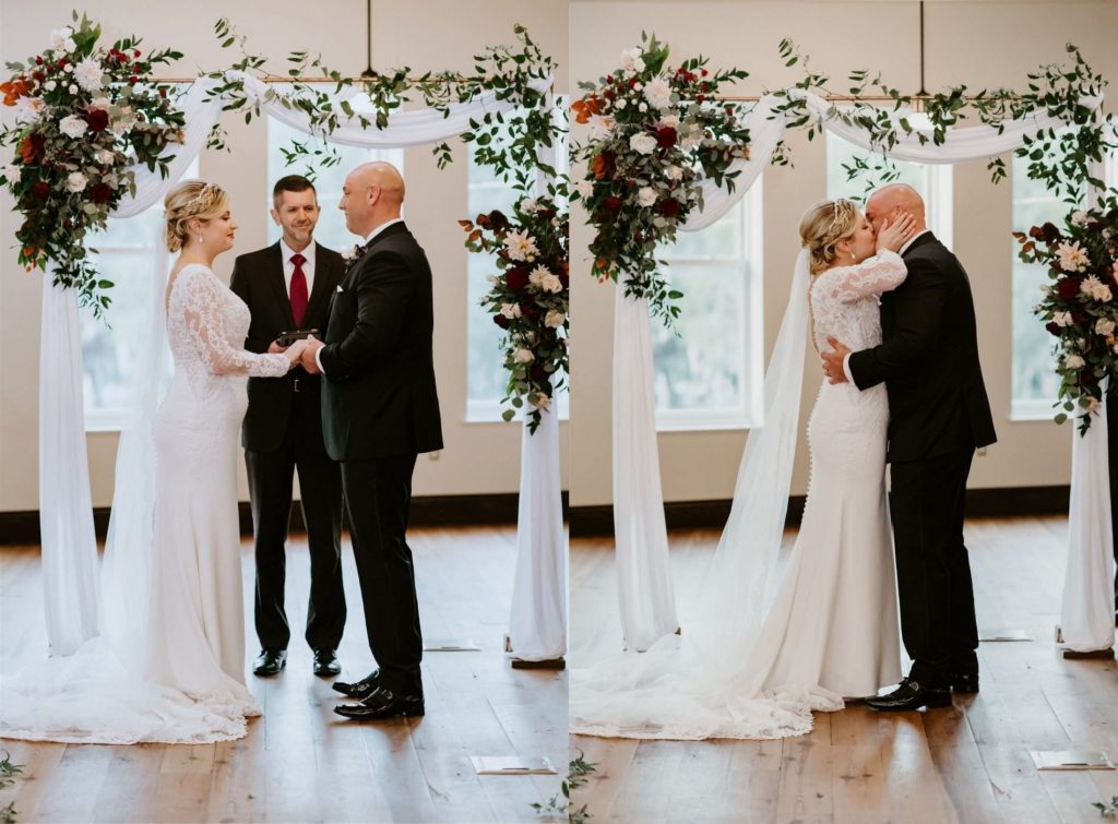 Bride and Groom First Kiss in front of Indoor Historic Industrial Loft Tampa Wedding Ceremony with Draped Arch Backdrop with Fall Greenery Floral Arrangement Spray of Copper Leaves, White and Red Roses, Zinnia, and Eucalyptus | Groom Wearing Classic Black Suit Tux | V Neck Long Sleeve Lace Sheath Bridal Gown with Long Cathedral Lace Veil