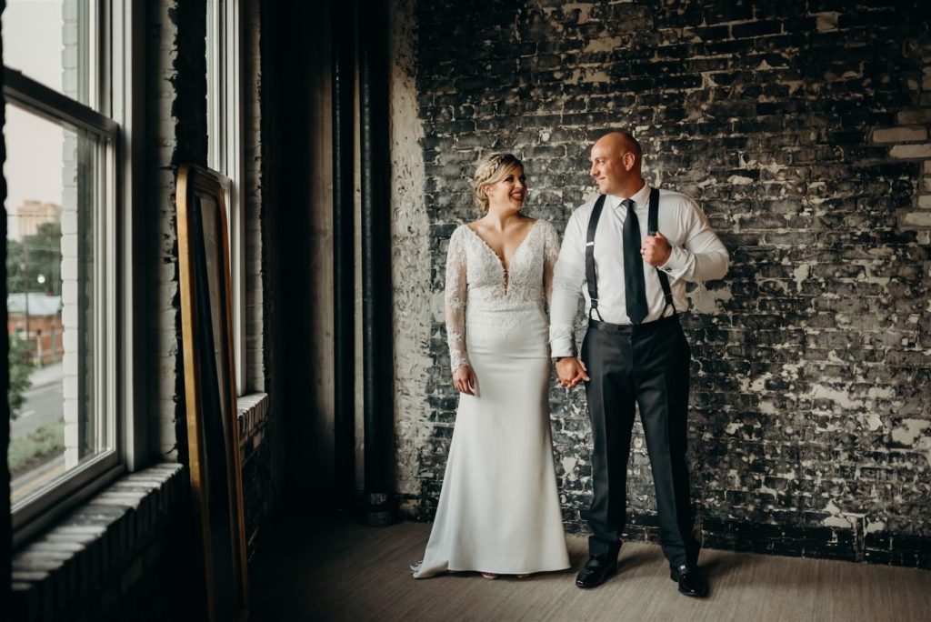 Indoor Bride and Groom Portrait at Industrial Loft Wedding with Exposed Brick Wall Backdrop | Groom Wearing Classic Black Suit Tux with Suspenders | V Neck Long Sleeve Lace Sheath Bridal Gown