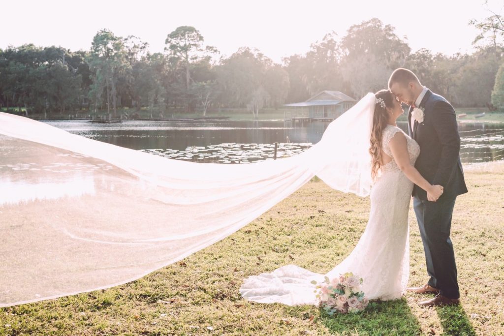 Romantic Lake Waterfront Sunset Bride and Groom Embracing Photo with Bride Veil Blowing in Wind | Tampa Bay Wedding Photographer Bonnie Newman Creative | Wedding Dress Truly Forever Bridal | Wedding Venue The Barn at Crescent Lake