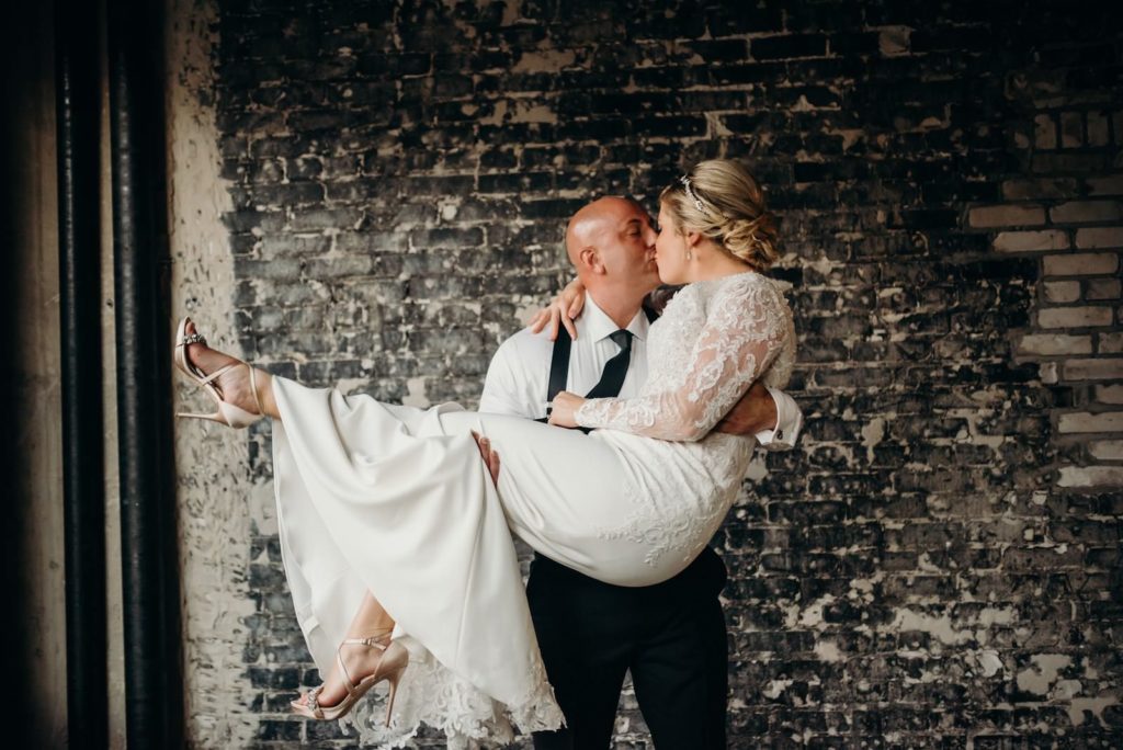Indoor Bride and Groom Kiss Portrait at Industrial Loft Wedding with Exposed Brick Wall Backdrop | Groom Wearing Classic Black Suit Tux with Suspenders | V Neck Long Sleeve Lace Sheath Bridal Gown