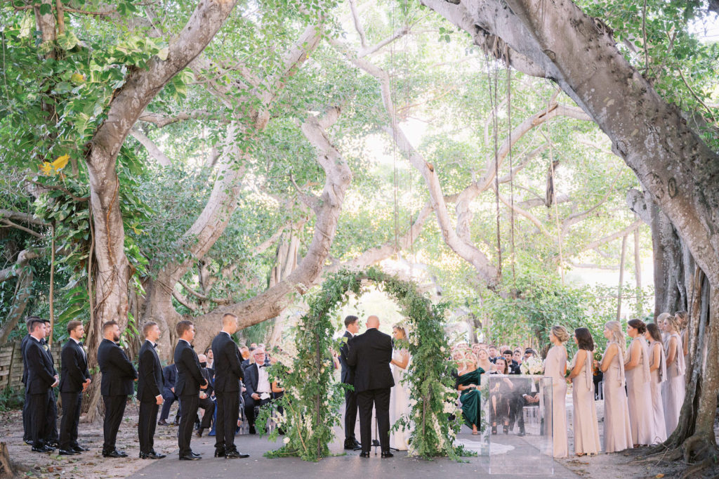 Outdoor Elegant Luxurious Wedding Ceremony Under Banyan Trees, Bride and Groom Exchanging Vows, Lush Greenery and White Floral Semi Circle Arch | Tampa Wedding Venue The Gasparilla Inn and Club