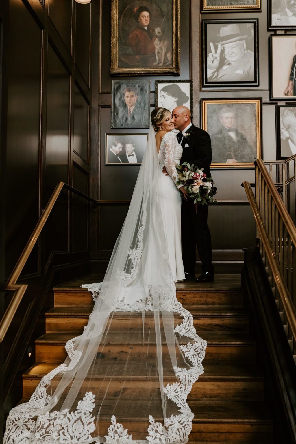 Indoor Bride and Groom Staircase Portrait at Industrial Loft Wedding | Groom Wearing Classic Black Suit Tux | V Neck Long Sleeve Lace Sheath Bridal Gown with Long Cathedral Lace Veil