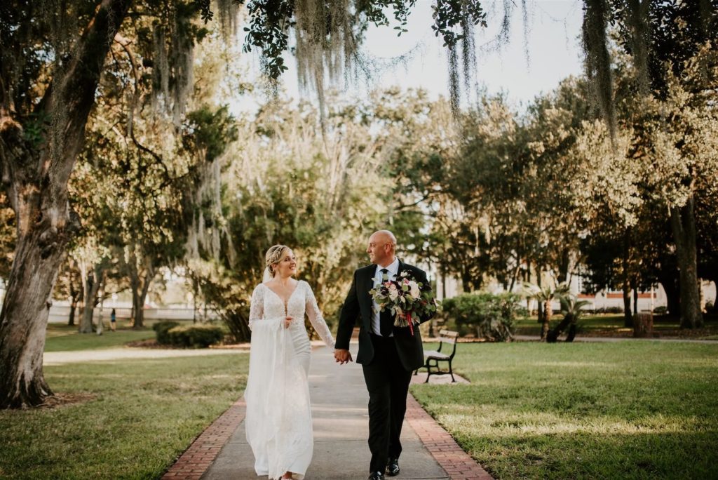 Outdoor Bride and Groom Downtown Tampa Portrait | Groom Wearing Classic Black Suit Tux | V Neck Long Sleeve Lace Sheath Bridal Gown with Long Cathedral Lace Veil