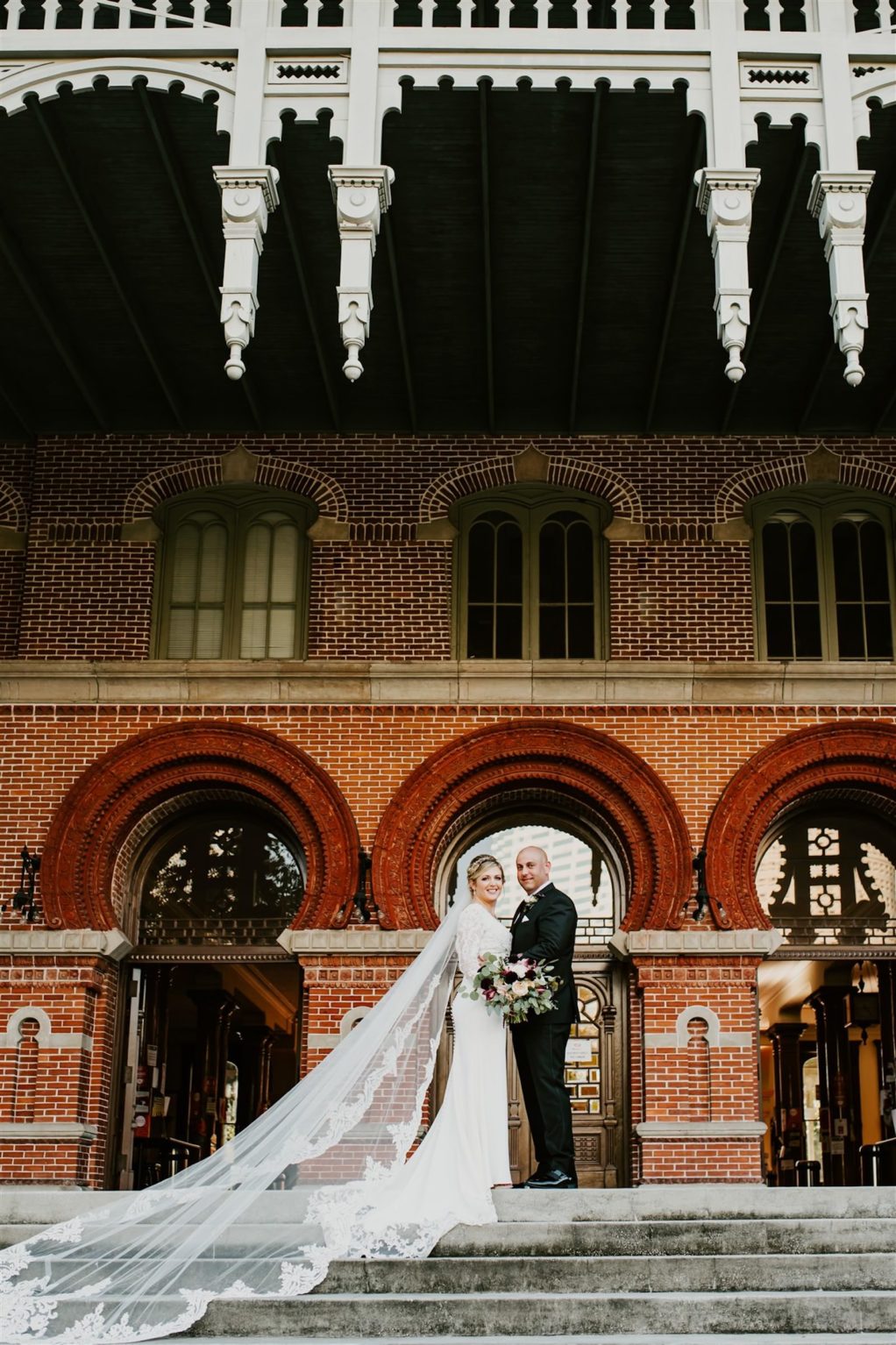 Outdoor Bride and Groom Downtown Tampa Portrait at UT Plant Museum | Groom Wearing Classic Black Suit Tux | V Neck Long Sleeve Lace Sheath Bridal Gown with Long Cathedral Lace Veil
