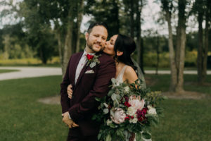 Romantic Dark and Moody Bride Holding Wild Garden Greenery Eucalyptus, Blush Pink King Proteas, White and Dark Red Floral Bouquet Kissing Groom Wearing Maroon Suit with Floral Printed Tie and Red Rose Boutonniere Romantic Outdoor Wedding Portrait | Tampa Bay Wedding Photographer Amber McWhorter Photography | Wedding Florist Brides & Blooms | Wedding Venue Paradise Spring