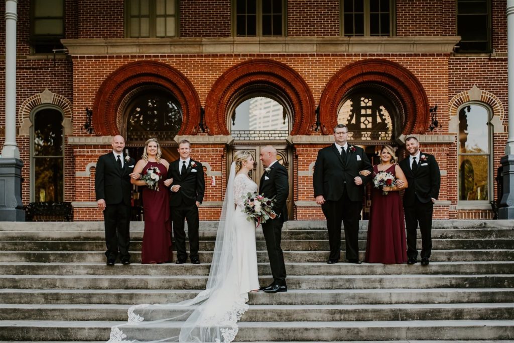 Outdoor Wedding Party Downtown Tampa Portrait at UT Plant Museum | Groom and Groomsmen Wearing Classic Black Suit Tux | V Neck Long Sleeve Lace Sheath Bridal Gown with Long Cathedral Lace Veil | Dark Red Burgundy Maroon Wine Long Bridesmaid Dresses