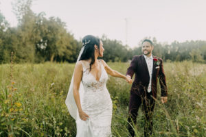 Romantic Bride Wearing Lace and Illusion Plunging V Neckline Wedding Dress Looking Back and Holding Hand of Groom in Tall Grass Wearing Maroon Suit with Floral Printed Tie | Tampa Bay Wedding Photographer Amber McWhorter Photography