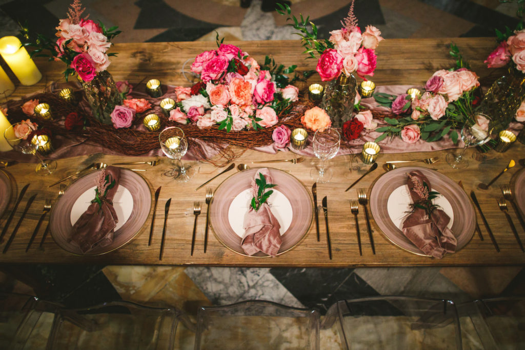 Luxury Elegant Tampa Wedding Reception with Long Wood Feasting Tables featuring Colorful Peach, Blush Pink, Fuchsia and Orange Rose Centerpieces with Greenery and Candles | Smoke Mauve Glass Charger Plates with Dusty Rose Velvet Napkins with Greenery Sprig and Gold Flatware Place Settings
