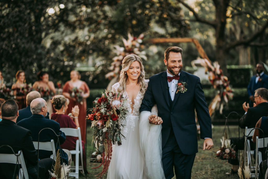 Rustic Florida Bride Holding Lush Floral Bouquet, King Proteas, Burgundy Flowers, Greenery and Red Hanging Amaranthus Walking Down the Wedding Ceremony Aisle After Exchanging Vows | Tampa Outdoor Wedding Venue Mision Lago Estate
