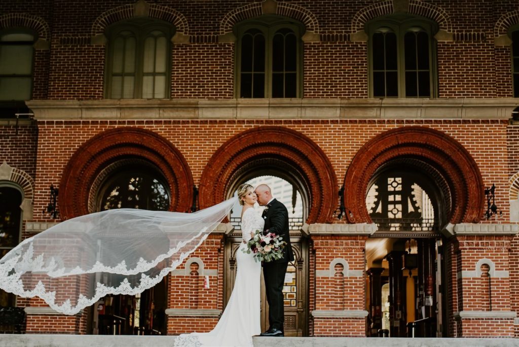 Veil Shot Wedding Photography | Outdoor Bride and Groom Downtown Tampa Portrait at UT Plant Museum | Groom Wearing Classic Black Suit Tux | V Neck Long Sleeve Lace Sheath Bridal Gown with Long Cathedral Lace Veil