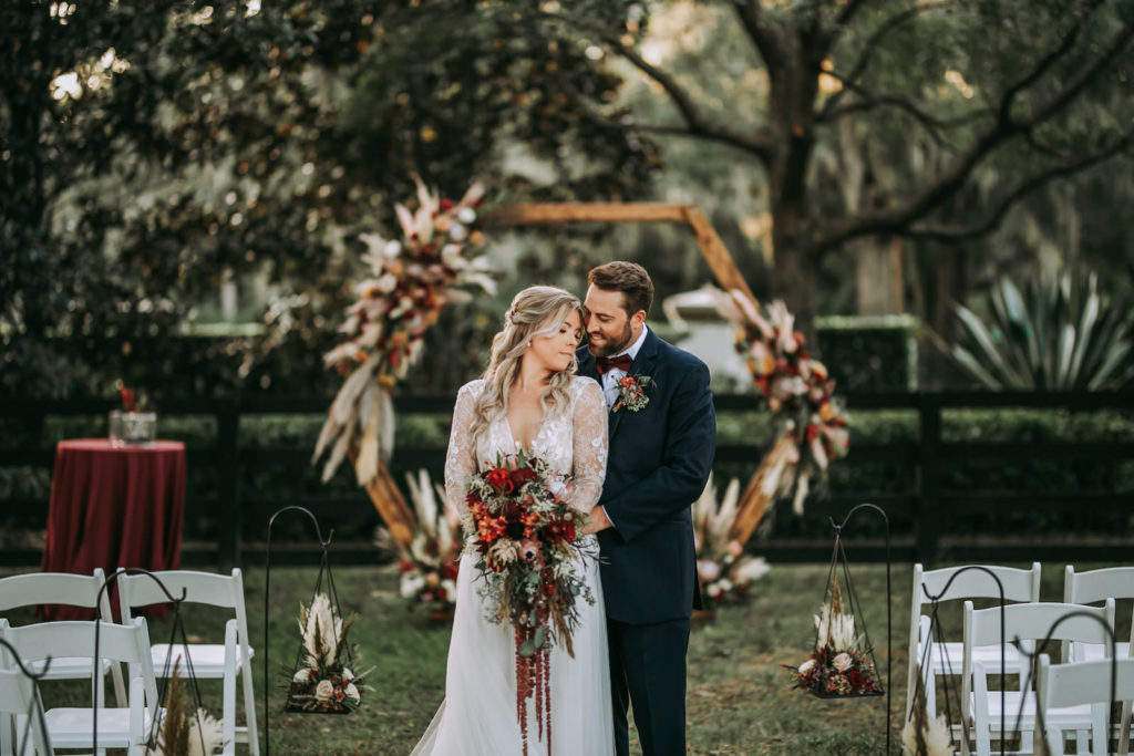 Rustic Florida Bride Holding Lush Floral Bouquet, King Protea, Greenery, Red Hanging Amaranthus, Burgundy and Burnt Orange Flowers and Groom Intimate Portrait | Outdoor Tampa Wedding Venue Mision Lago Estate