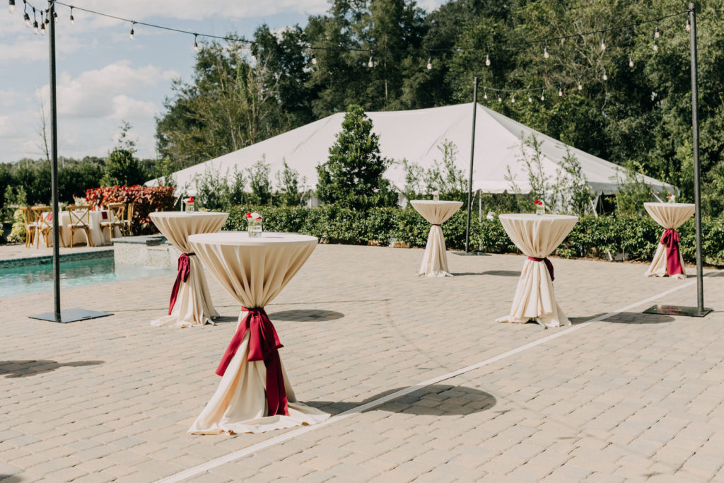 Romantic Courtyard Pool Cocktail House Decor, String Lights, High Round Tables with Ivory Linens Tied with Maroon Ribbons | Tampa Bay Wedding Photographer Amber McWhorter Photography | Wedding Venue The Secret Garden at Paradise Spring