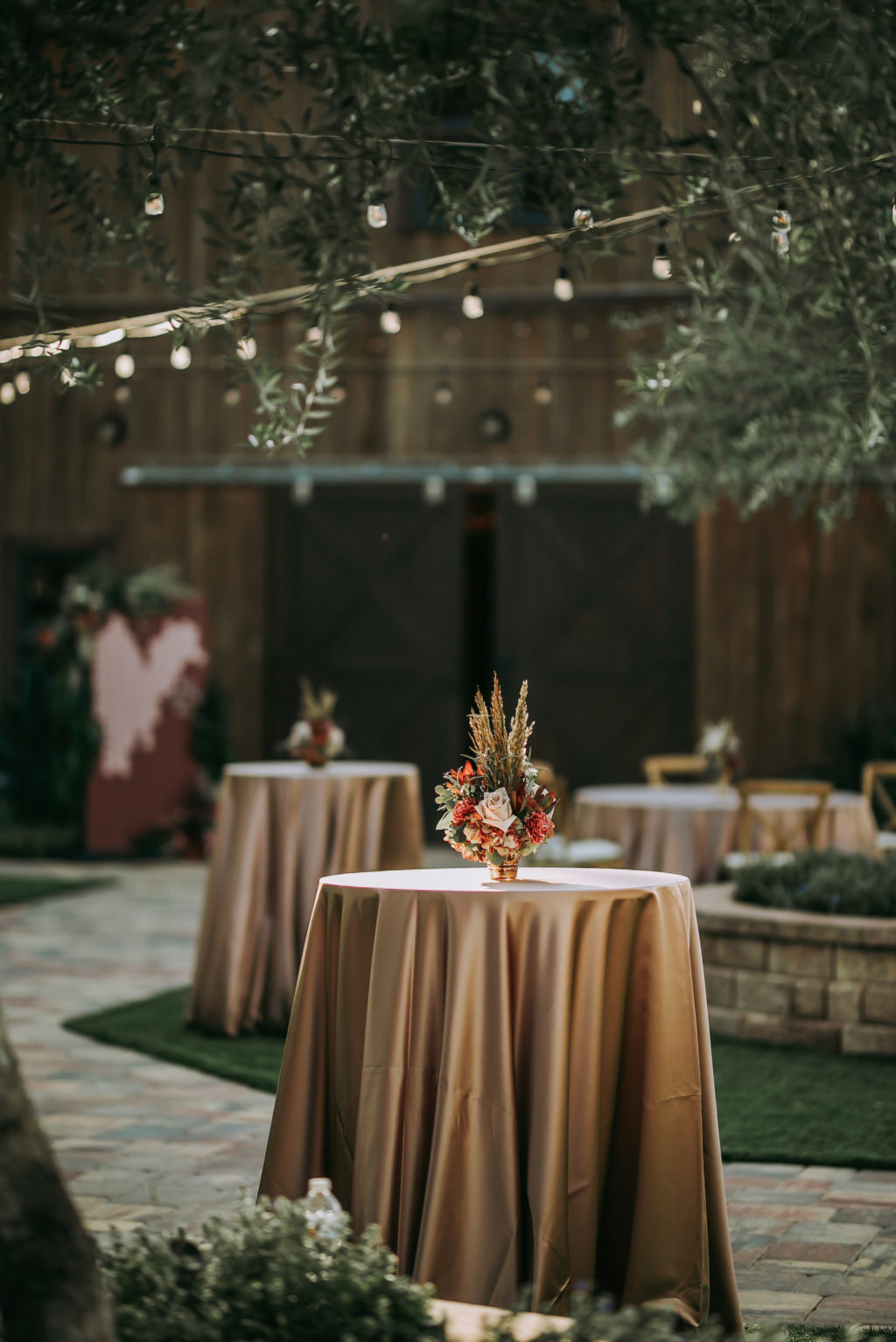Outdoor Rustic Cocktail Hour Wedding Decor, String Lights, Tall Tables with Champagne/Gold Table Linens, Small Floral Centerpiece with Pampas Grass