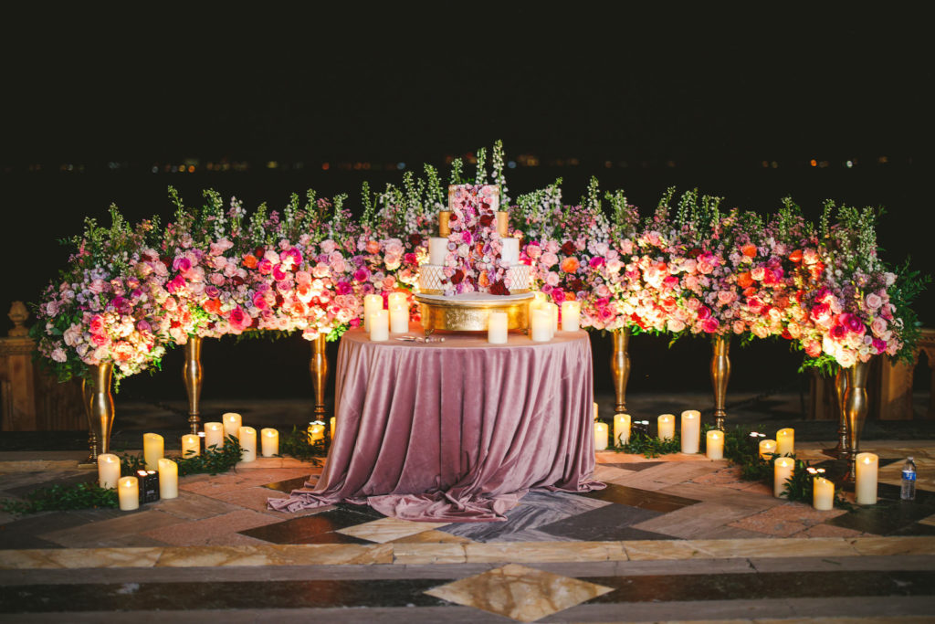 Outdoor Waterfront Tampa Bay Sarasota Wedding Cake Display with Ground Arch Floral Arrangements featuring Colorful Pink Purple and Orange Roses with Candles | Mauve Dusty Rose Velvet Cake Table Linen with Gold Ornate Round Cake Stand | Four Tier White and Metallic Gold Luxury Wedding Cake by The Artistic Whisk featuring Fresh Flower Roses