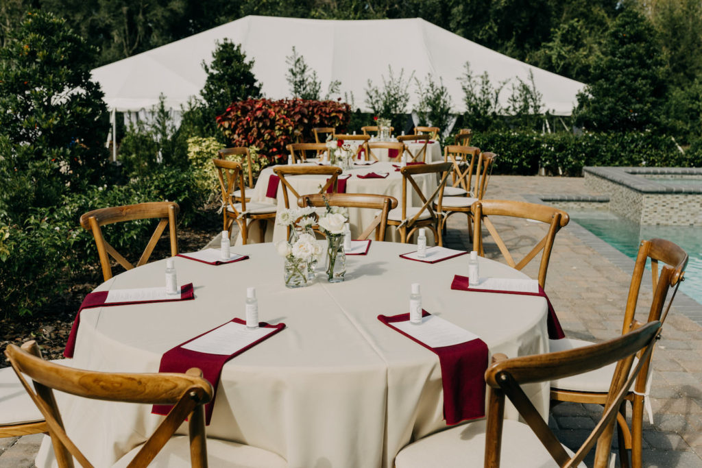 Outdoor Courtyard Pool Wedding Reception Decor, Round Tables with Ivory Tablecloth, Maroon Linen Napkins, Low White Roses Floral Centerpiece, Wooden Crossback Chairs | Tampa Bay Wedding Photographer Amber McWhorter Photography | Wedding Venue Paradise Spring