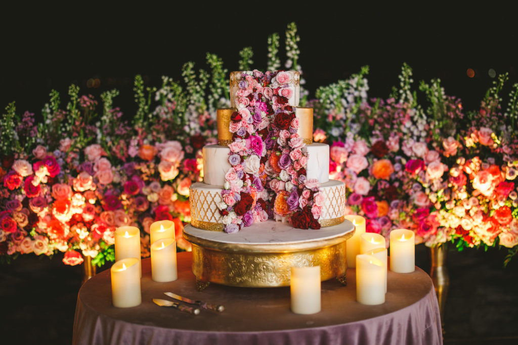 Outdoor Waterfront Tampa Bay Sarasota Wedding Cake Display with Ground Arch Floral Arrangements featuring Colorful Pink Purple and Orange Roses with Candles | Mauve Dusty Rose Velvet Cake Table Linen with Gold Ornate Round Cake Stand | Four Tier White and Metallic Gold Luxury Wedding Cake by The Artistic Whisk featuring Fresh Flower Roses