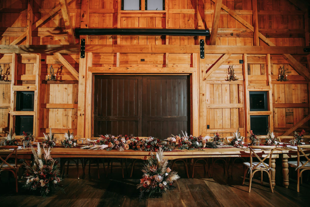 Rustic Barn Wedding Reception, Long Feasting Table for the Wedding Party with Lush Floral Arrangements, Pampas Grass, King Proteas, Orange and Burgundy Roses, Wooden Crossback Chairs | Tampa Wedding Venue Mision Lago Estate | Mision Lago Ranch
