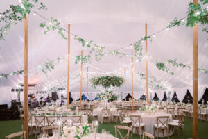 Luxurious Elegant Classic Florida Outdoor Tent Wedding Reception Decor, White Linen Draping, String Lights with Greenery Vines and a Lush Greenery Leaves Wreath Chandelier, Round and Long Tables with Taupe Tablecloths, Wooden Cross Back Chairs, Candlesticks in Glass Hurricane Tumblers