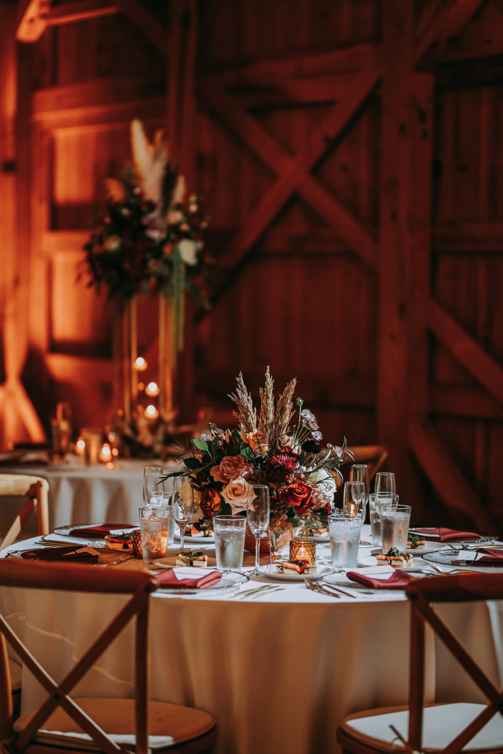 Rustic Barn Wedding Reception Decor, Round Table with Ivory Linen, Wooden Cross Back Chairs, Low Floral Centerpiece with Orange and Burgundy Roses, Pampas Grass