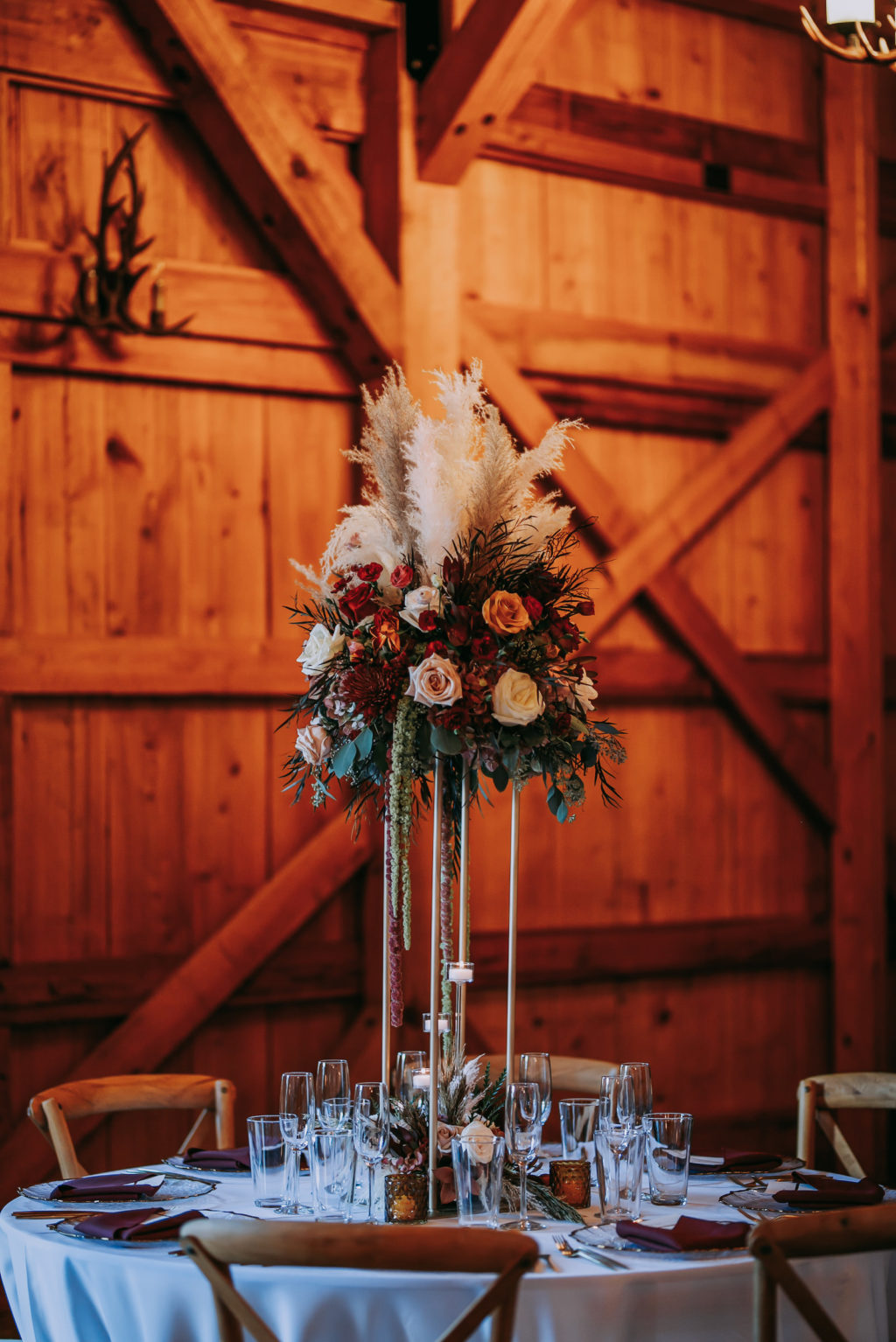 Rustic Barn Wedding Reception Decor, Tall Geometric Stand with Lush Floral Centerpiece, Pampas Grass, Hanging Amaranthus, Blush and Orange Roses, Burgundy Flowers, Wooden Cross Back Chairs
