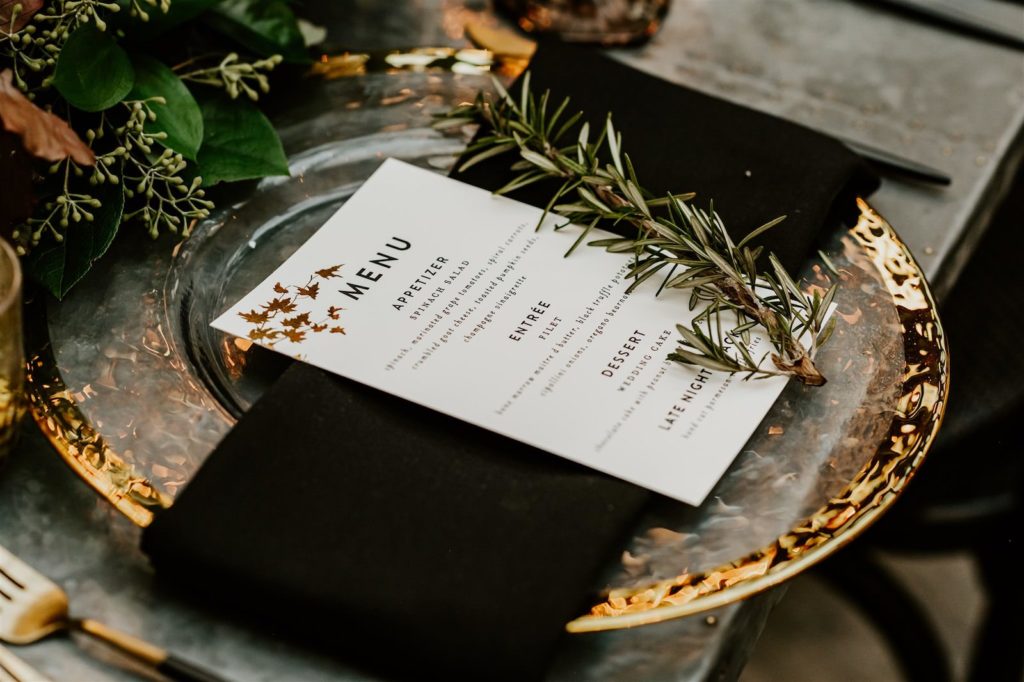Wedding Place Settings with Gold Rim Glass Charger Plates, Black and Gold Flatware, Black Napkins, Menu Cards, and Sprig of Rosemary by Kate Ryan Event Rentals