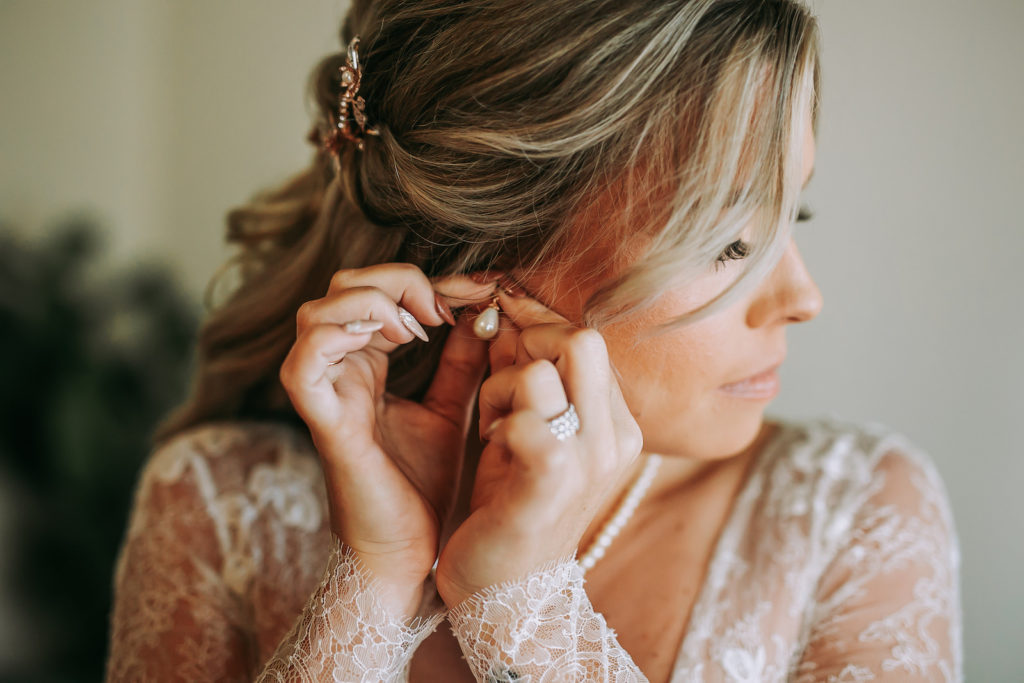 Bride Getting Wedding Ready Putting on Pearl Earring in Lace and Illusion Long Sleeve Wedding Dress | Michele Renee the Studio