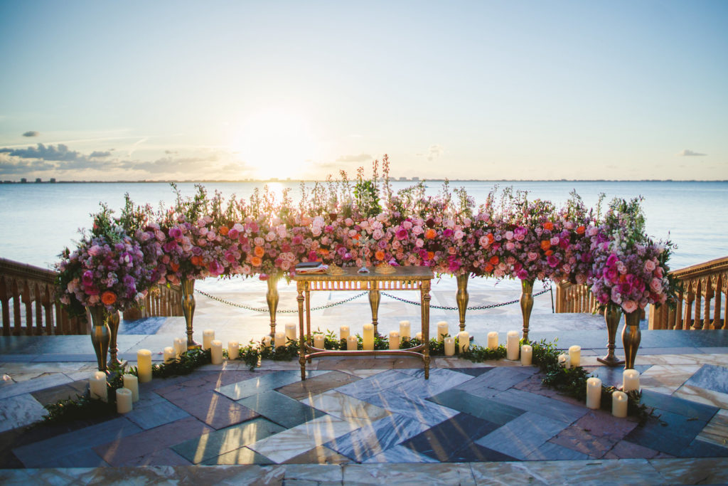 Outdoor Waterfront Tampa Bay Sarasota Wedding Ceremony with Ground Arch Floral Arrangements featuring Colorful Pink Purple and Orange Roses with Candles | Sarasota Luxury Wedding