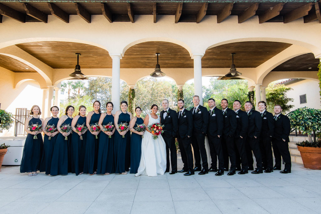 Bride Holding Colorful Jewel Toned Floral Bouquet, Groom in Black Tuxedo, Bridesmaids in Matching Navy Blue Halter Dresses, Groomsmen in Black Suits