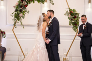 Florida Bride and Groom Exchanging Wedding Ceremony First Kiss Under Gold Geometric Hexagonal Arch with Greenery and Blush Pink, Burgundy Roses | Tampa Bay Wedding Planner Coastal Coordinating