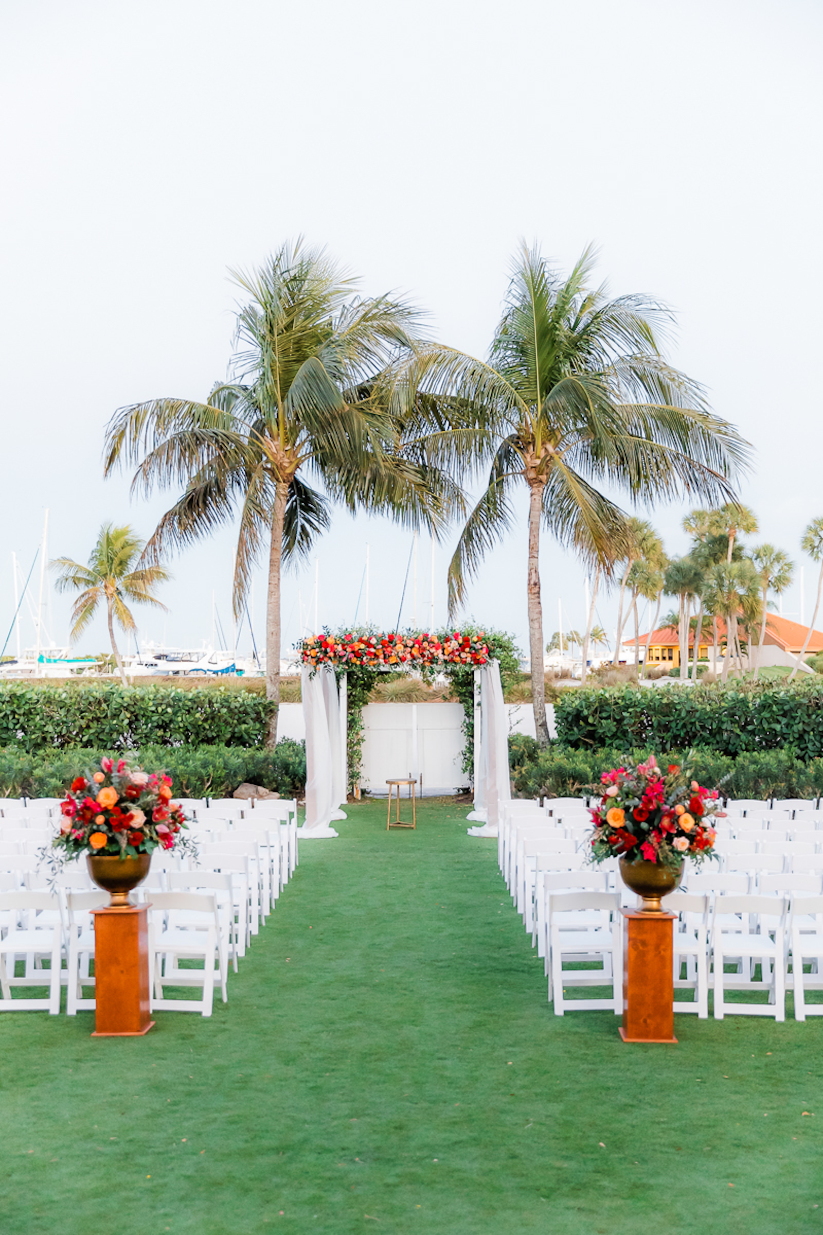 Elegant Waterfront Outdoor Garden Wedding Ceremony Decor, White Folding Chairs, Tall Wooden Aisle Pedestals with Jewel Tone Floral Arrangements, Arch with White Linens and Flowers | Tampa Bay Wedding Venue The Resort at Longboat Key Club