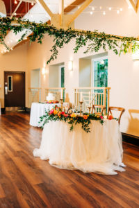 Garden Style Wedding Reception Décor Inspiration | White Linen and Circular Table with Wooden Chairs and Hanging Greenery | Vibrant Pink, Yellow, and Orange Centerpieces | Sweetheart Table Ideas