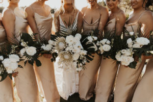Neutral Timeless Bride with Bridesmaids in Silky Matching Champagne Gold Dresses Holding Floral Bouquets with White Roses, King Proteas, Monstera Palm Leaves, White Orchids and Palm Fronds
