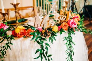 Sweetheart Table with Wooden Chairs and Hanging Greenery | Vibrant Pink, Yellow, and Orange Rose Centerpieces | Garden Style Wedding Reception Décor Ideas