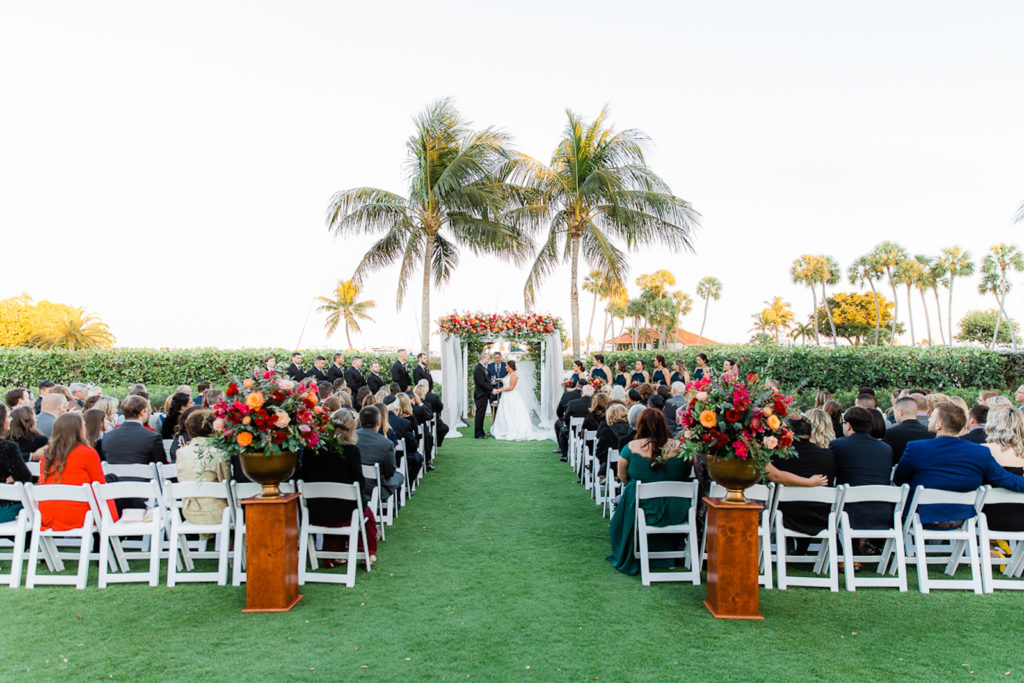Elegant Waterfront Outdoor Garden Wedding Ceremony Decor, Bride and Groom Exchanging Wedding Vows, White Folding Chairs, Tall Wooden Aisle Pedestals with Jewel Tone Floral Arrangements, Arch with White Linens and Flowers | Tampa Bay Wedding Venue The Resort at Longboat Key Club