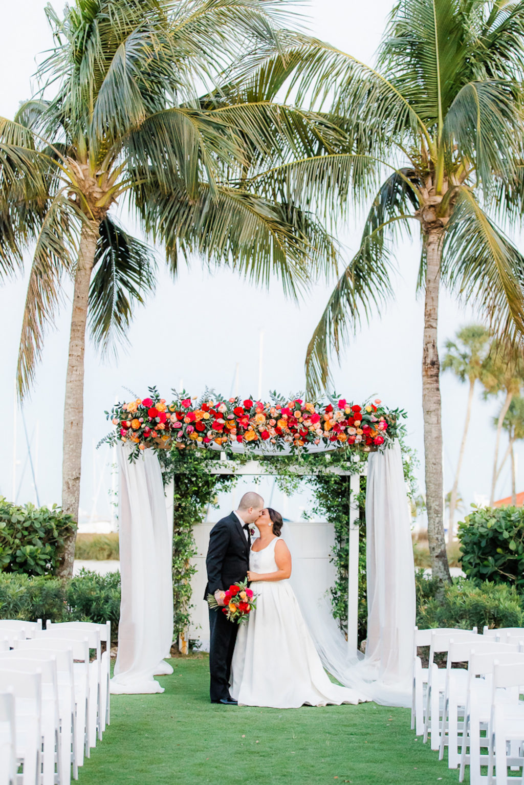 Florida Bride and Groom Kissing Under Wedding Ceremony Arch with White Linens with Colorful Jewel Tone Pink, Orange, Red Flowers and Greenery | Tampa Outdoor Waterfront Garden Wedding Venue The Resort at Longboat Key Club