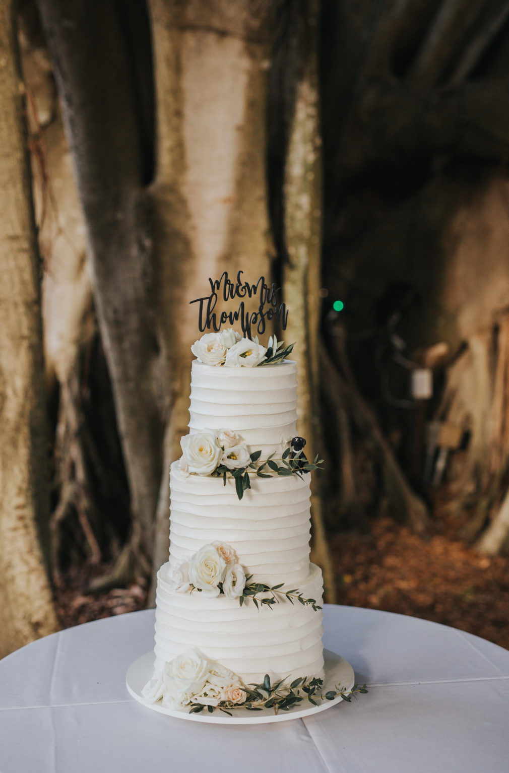 Rustic Three Tiered Round Frosted Icing Wedding Cake, White Flower Details and Greenery, and Black Mr. & Mrs. Cake Topper